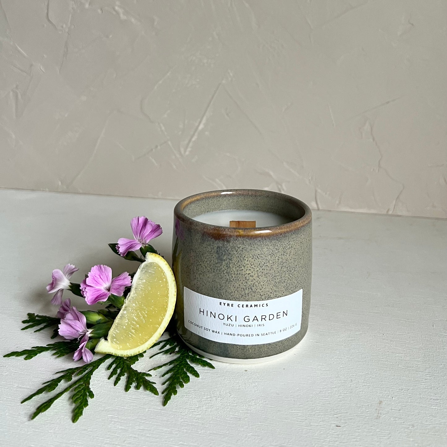 Ceramic candle labeled 'Hinoki Garden' with a slice of lemon and purple flowers on a textured surface.