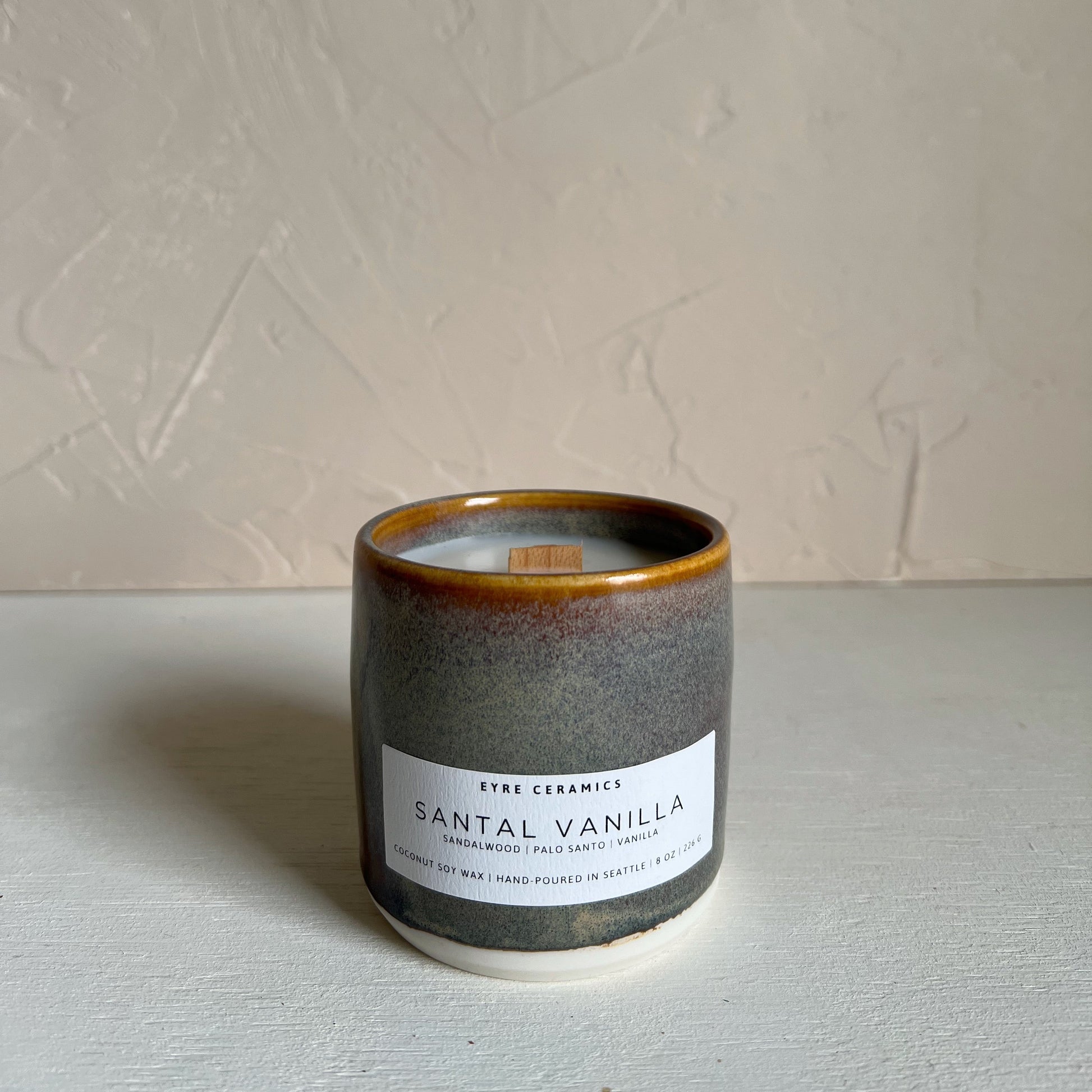 Candle in a ceramic holder labeled 'Santal Vanilla' on a neutral background