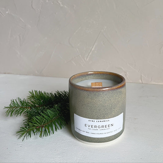 Ceramic candle labeled 'Evergreen' with a small branch on a light surface and light gray background