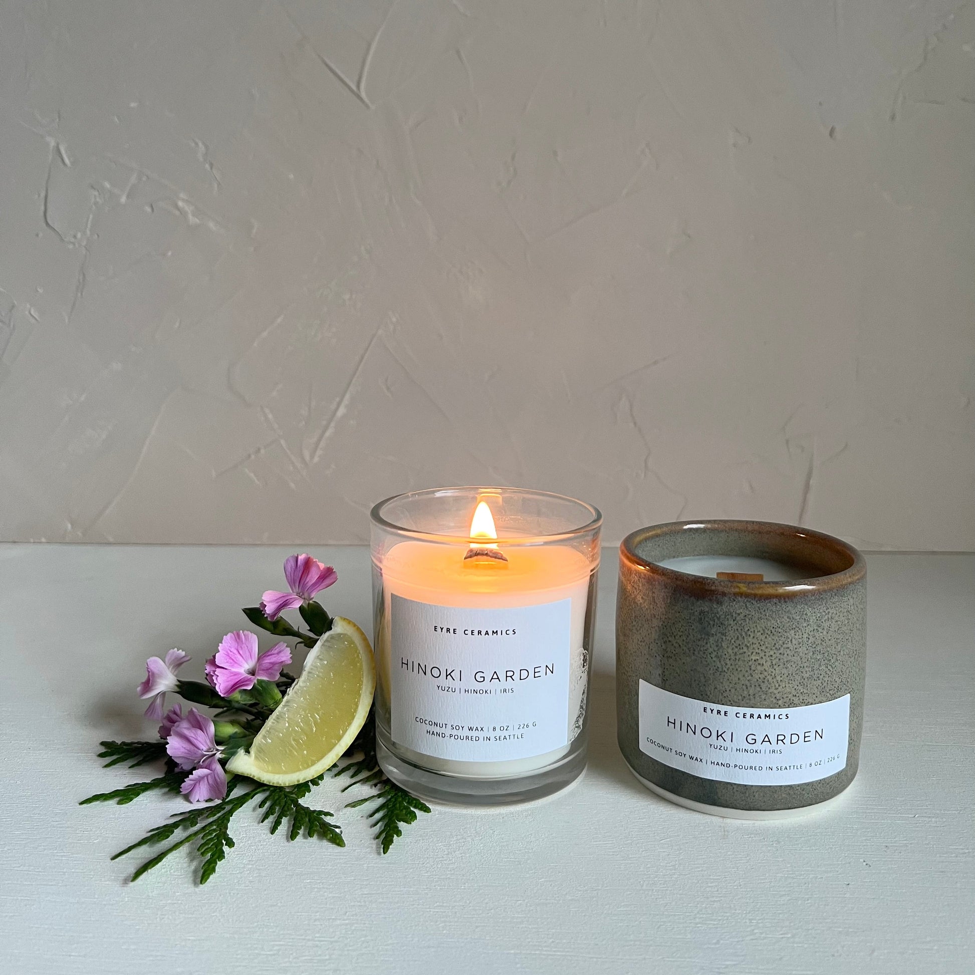 Two candles from Hinoki Garden on a white surface with flowers and a lime wedge.