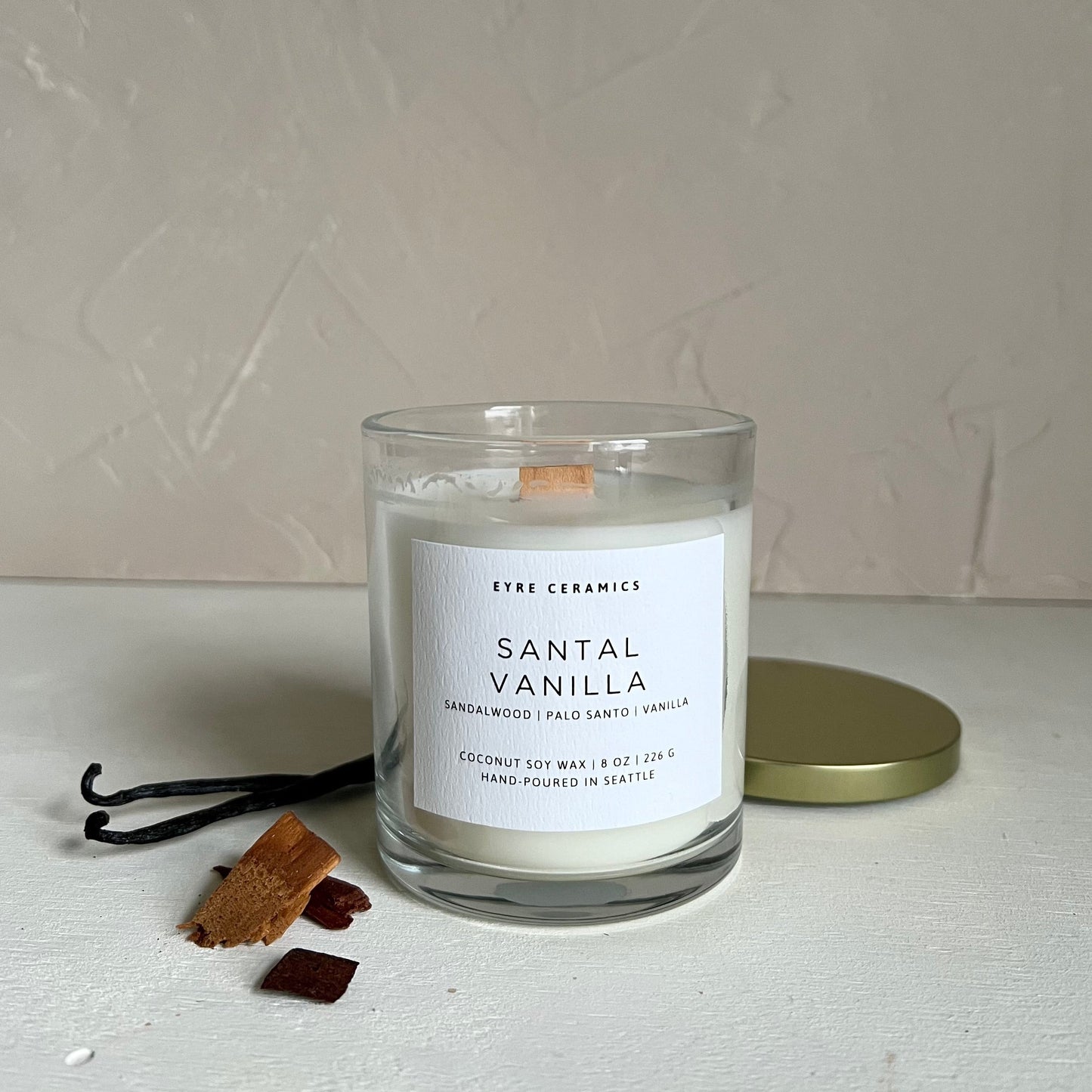 Candle labeled 'Santal Vanilla' with vanilla beans and a sandalwood on a neutral background