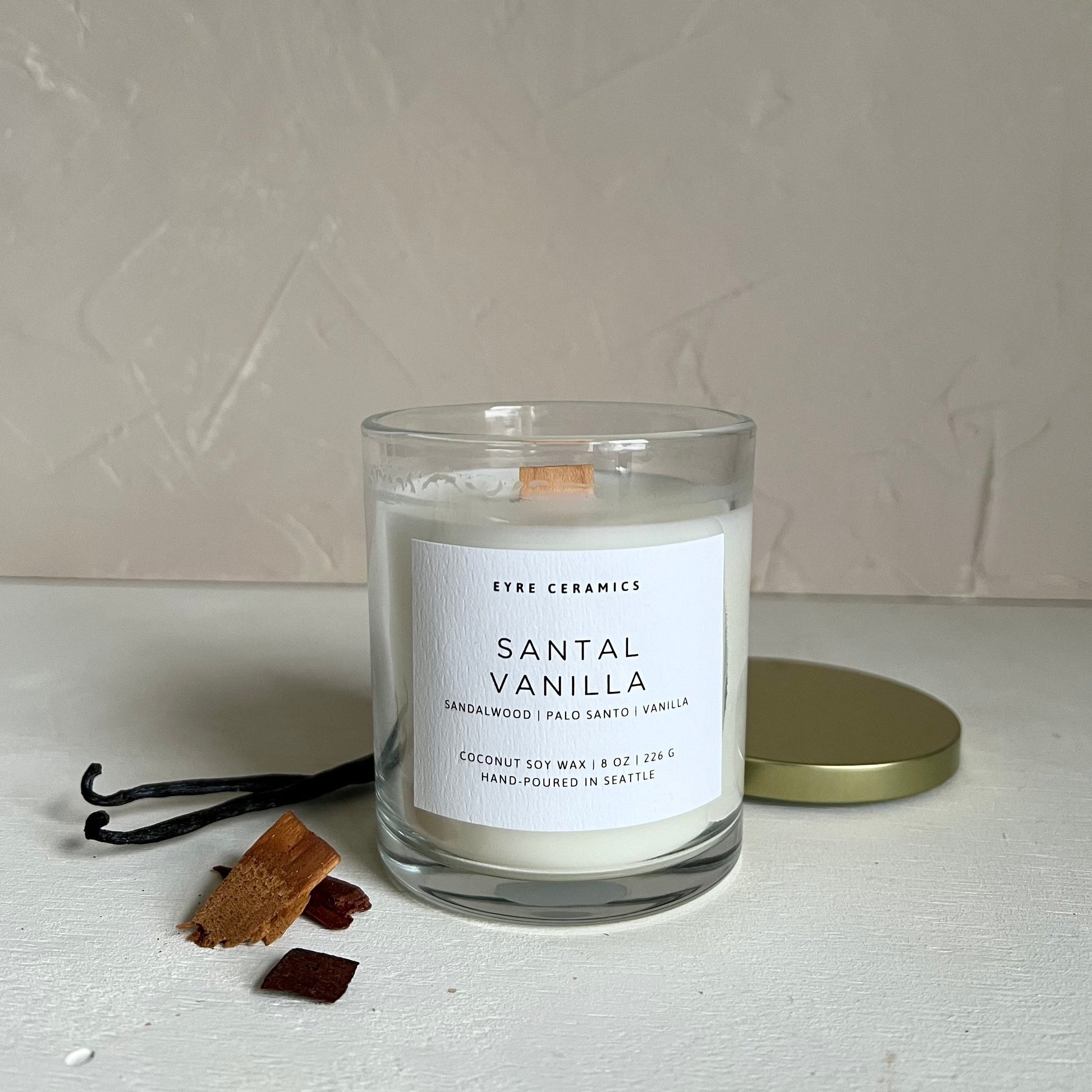 Candle labeled 'Santal Vanilla' with vanilla beans and a sandalwood on a neutral background