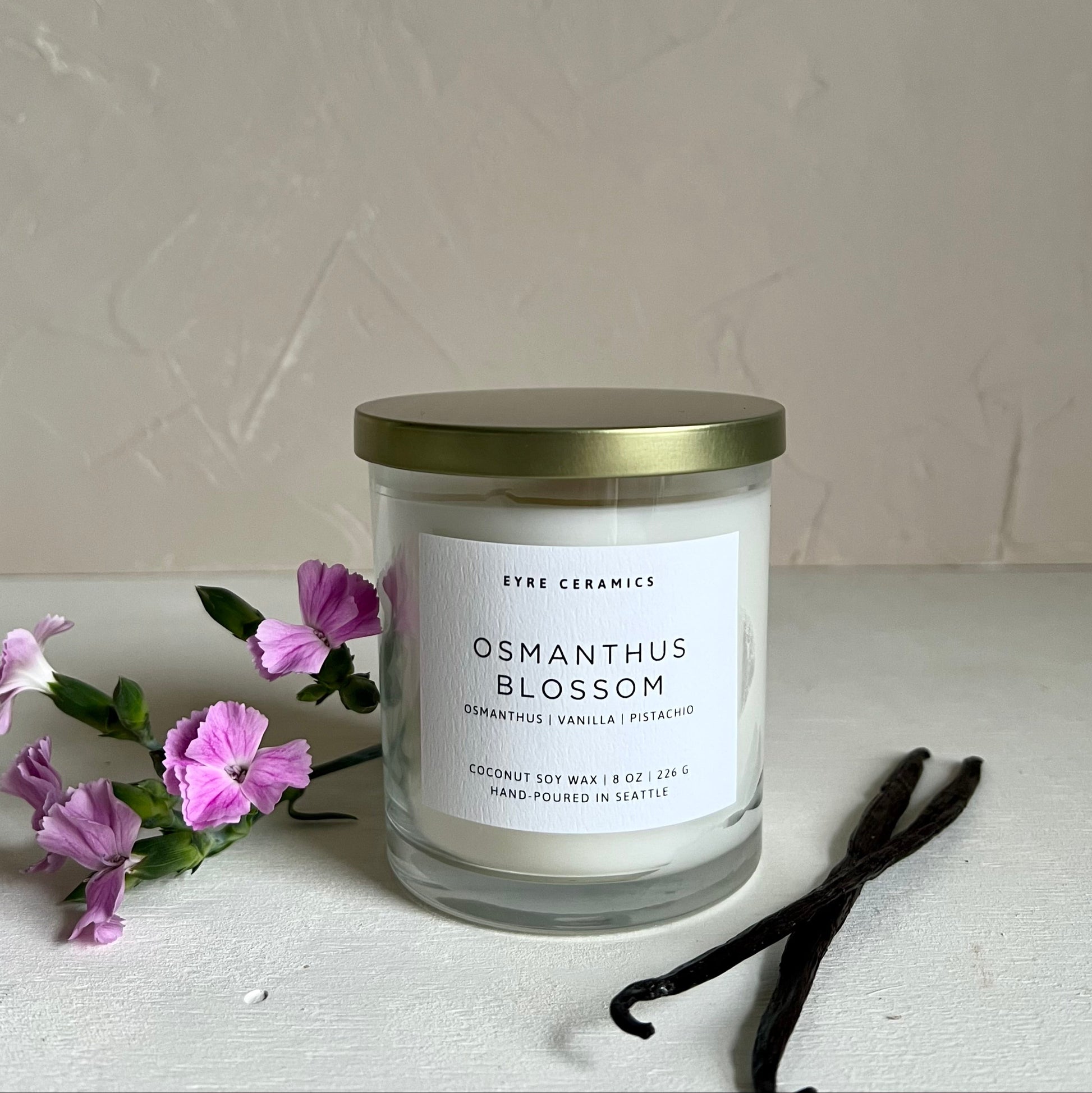 Candle labeled 'Osmanthus Blossom' with a gold lid on a white surface with purple flowers and vanilla beans.