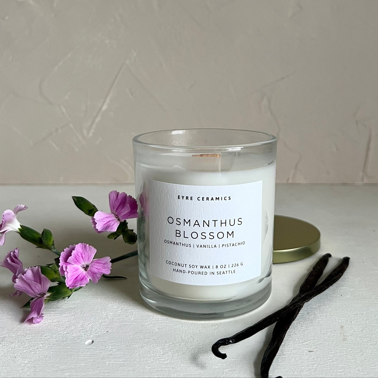 Candle labeled 'Osmanthus Blossom' with purple flowers and vanilla beans on a light surface.