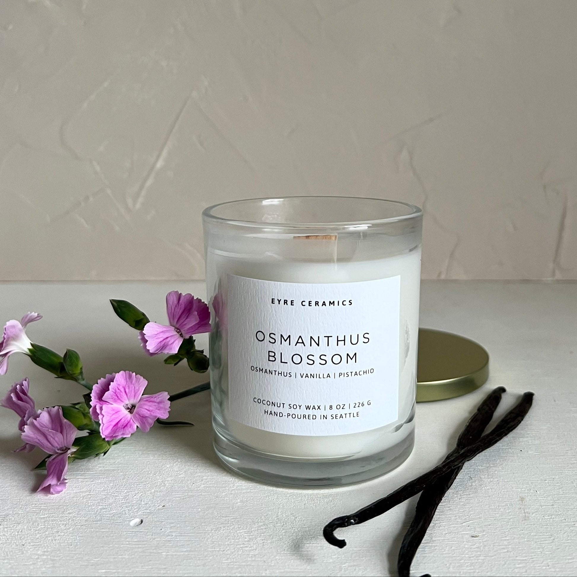Candle labeled 'Osmanthus Blossom' with purple flowers and vanilla beans on a light surface.