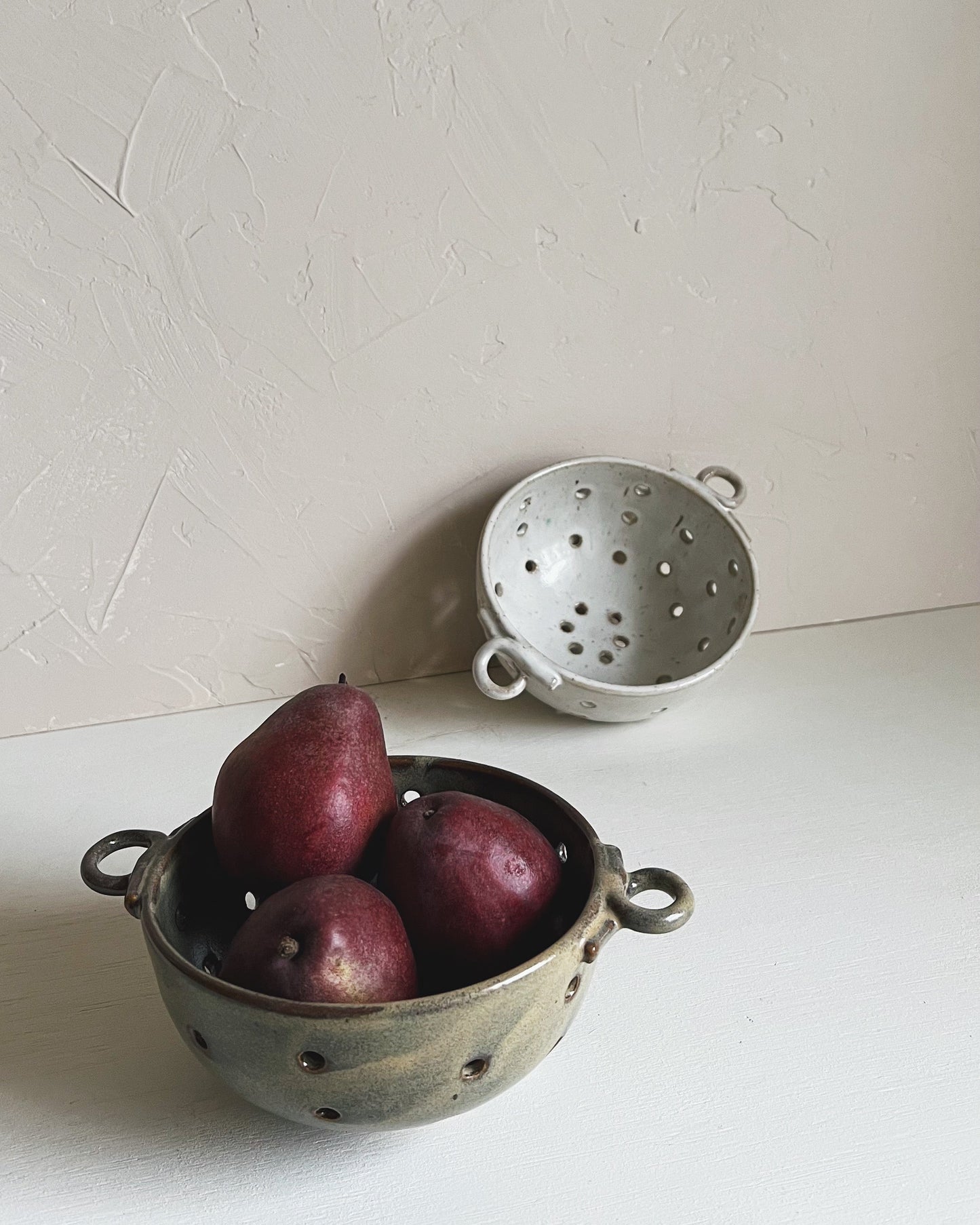 Two ceramic berry bowl colanders with red pears on a white surface and light gray background