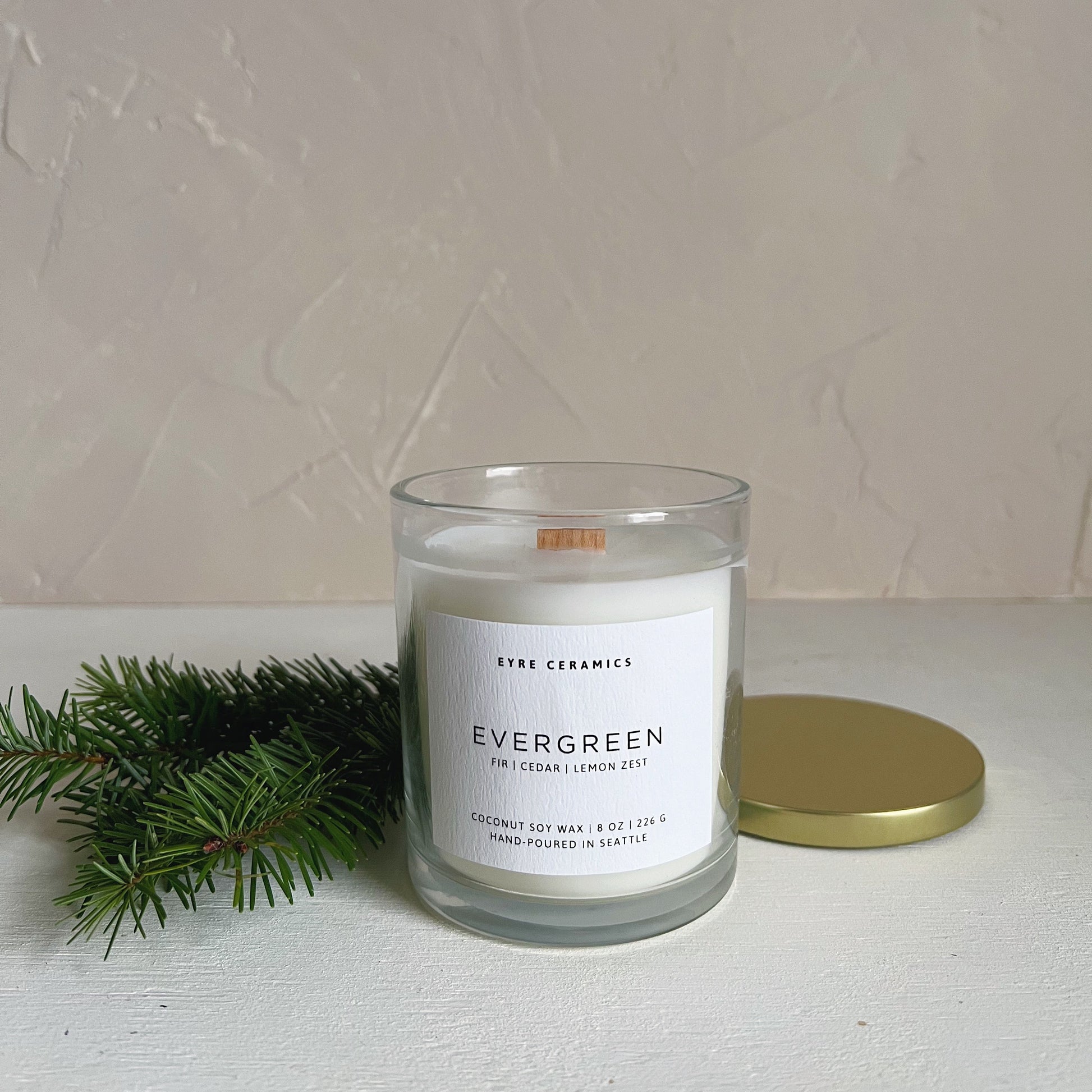 Candle labeled 'Evergreen' with a gold lid and greenery on a white surface.