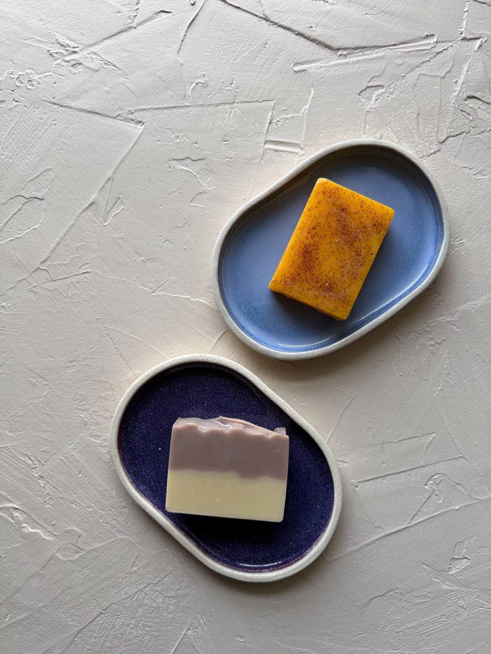 Two soap bars on blue ceramic trays against a textured gray background